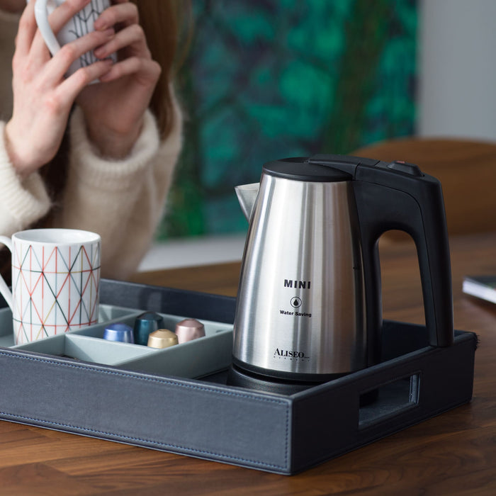 ALISEO Mini Wasserkocher steht auf einem Tablett mit Tassen. Das Tablett steht auf einem Holztisch vor einer Person, die ihren Kaffee oder Tee aus einer Tasse trinkt.