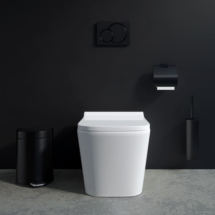 Schwarze ALISEO Bad-Accessoires. WC-Bürstengarnitur, Rollenhalter und Abfalleimer in Schwarz matt. In einem schlichten Badezimmer mit einer schwarzen Wand und weißem WC.